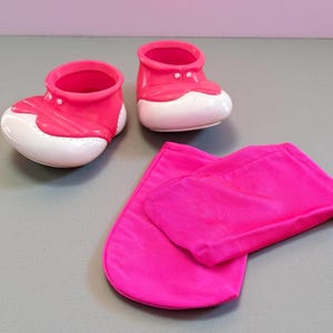 Może przedstawiać: Para jasnoróżowych bucików dziecięcych z białymi podeszwami i pasującym różowym zestawem kocyków. Buty mają zaokrąglony kształt i są wykonane z błyszczącego materiału. Zestaw kocyków jest złożony i ma miękką fakturę.