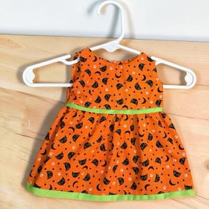 Op de afbeelding: Een mouwloze jurk in feloranje met een herhalend patroon van zwarte kattengezichten, maansikkels en witte pootafdrukken. De jurk is voorzien van een limoengroene horizontale band in de taille en een bijpassende limoengroene bies langs de onderzoom.