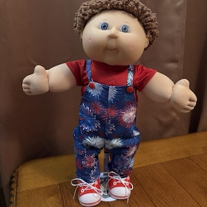 May include: A doll with a round face, blue eyes, and brown curly hair wearing a red short-sleeved shirt under navy blue overalls featuring a red and white firework pattern. The doll wears red and white trainers with white laces.