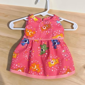 Può includere: Un vestito rosa senza maniche con un motivo ripetuto di orsetti colorati dei cartoni animati, cuori bianchi e stelle gialle. L'abito presenta una vita arricciata, uno scollo rotondo e un bordo dell'orlo rosa tinta unita, su una gruccia bianca.