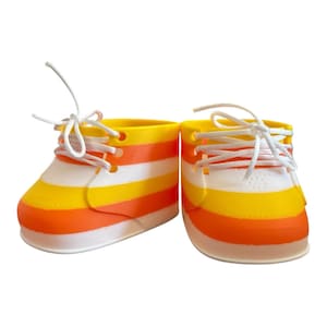 May include: A pair of colorful baby shoes with a candy corn design. The shoes feature horizontal stripes of white, orange, and yellow. White laces are tied at the top. The shoes are designed for a small child.