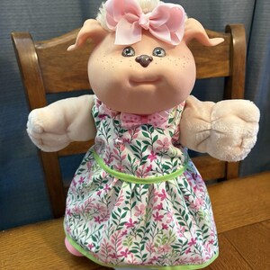 Puede incluir: Una muñeca de peluche con cara de vinilo, pecas y orejas caídas, que lleva un lazo rosa claro en la cabeza. La muñeca viste un vestido blanco sin mangas con un estampado floral verde y rosa, un pequeño lazo rosa en el cuello y un cascabel.