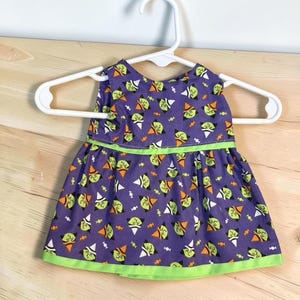 May include: A sleeveless dress featuring a purple background with a repeating pattern of green-faced witches wearing orange and black hats, interspersed with small white candies. The dress has a bright green trim at the waist and the hemline.