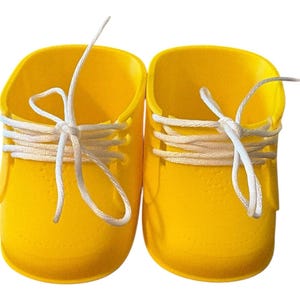 May include: A pair of bright yellow baby shoes with white laces tied in bows. The shoes have a rounded toe and a textured surface. The laces are threaded through eyelets, creating a classic sneaker look.