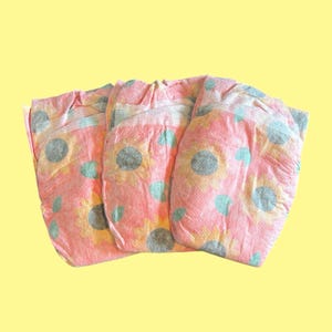 Puede incluir: Tres pañales rosas para bebés con un estampado de girasoles y lunares azules están dispuestos sobre un fondo amarillo. Los pañales tienen un aspecto suave y texturizado, lo que sugiere comodidad y absorción. El diseño es alegre y adecuado para bebés.