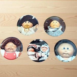 Könnte beinhalten: Eine Sammlung von fünf runden Knöpfen mit Bildern von Cabbage Patch Kids-Puppen. Die Knöpfe zeigen verschiedene Puppen mit unterschiedlichen Frisuren, Outfits und Gesichtsausdrücken. Die Knöpfe sind vor einem hellen Holzmaserungshintergrund angeordnet.