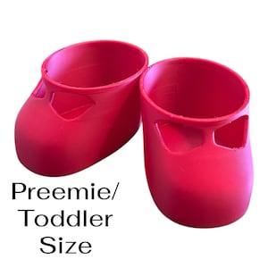 May include: Two bright red preemie/toddler-sized shoes. The shoes are made of a solid material and have a rounded toe and a small cutout on the side. The text "Preemie/Toddler Size" is printed in black below the shoes.