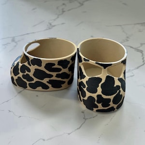 May include: Two ceramic planters with a beige base and black spotted pattern. One planter has a handle-like cutout, while the other has two rectangular cutouts. The interior of both planters is beige.