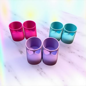 May include: Six colorful, small, cylindrical containers. Two are bright pink, two are light blue, and two are lavender. Each has a bow-shaped cutout near the top. The containers are arranged on a white and gray marble-like surface.