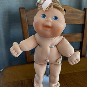May include: A vintage Cabbage Patch Kid doll with blonde yarn hair, blue eyes, and a pink star on its cheek. The doll is nude, with a soft, flesh-toned body and a small tag attached. It stands on a white stand.