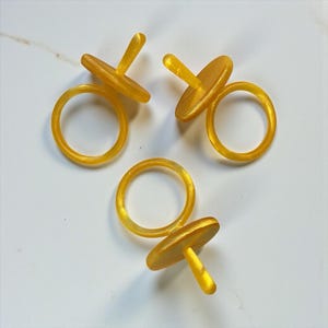 Puede incluir: Tres anillos amarillos en forma de chupete sobre una superficie blanca. Cada anillo tiene una base circular y una parte superior plana y redonda con un pequeño mango recto. Los anillos están hechos de un material translúcido y brillante.