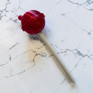 May include: A large, red lollipop with a wooden stick. The candy is a vibrant red color and has a round shape with a small, circular detail on top. The stick is a light beige color.