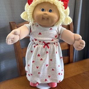 Boneca Cabbage Patch Kid dos anos 80, cabelo amarelo-limão, olhos azuis, um dentinho, vestido com corações e maria-chiquinhas.
