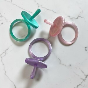 May include: Three resin rings in teal, pink, and purple, each with a pacifier-shaped top. The rings are displayed on a white marble surface, highlighting their glossy finish and unique design. The pacifier tops have a circular shape.