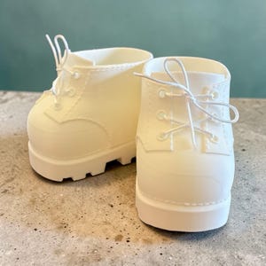 Könnte beinhalten: Zwei cremeweiße Miniaturstiefel mit Schnürsenkeln. Die Stiefel haben ein klassisches Design mit runder Spitze, Schnürverschluss und klobiger Sohle. Die Stiefel bestehen aus einem festen Material und stehen vor einem neutralen Hintergrund.