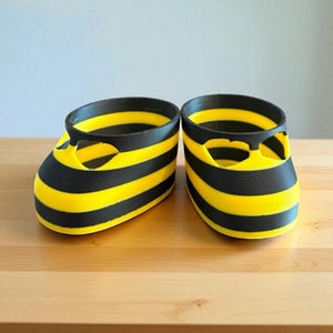 Puede incluir: Un par de zapatos de bebé novedosos con un diseño de abeja. Los zapatos son amarillos con rayas horizontales negras y un ribete negro alrededor de la parte superior. Los zapatos están sobre una superficie de madera.