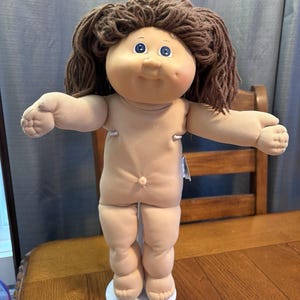 Puede incluir: Una muñeca Cabbage Patch Kid vintage con cabello de hilo marrón y ojos azules. La muñeca tiene un cuerpo suave color carne y está de pie sobre una base blanca. Los brazos de la muñeca están extendidos.