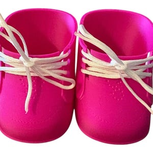 May include: A pair of vibrant pink baby shoes with white laces. The shoes have a textured finish and a rounded toe design. The laces are tied in a bow. The shoes are set against a white background.