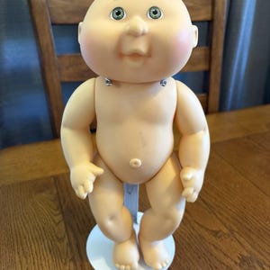 May include: A vintage Cabbage Patch Kid doll with a round, pale skin head, rosy cheeks, and green eyes. The doll is nude and stands on a white base. The doll's arms and legs are slightly bent.