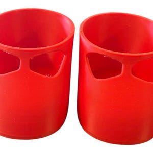 Puede incluir: Dos objetos cilíndricos de color rojo brillante, con base redondeada y un par de recortes en forma de ojo cerca de la parte superior. Los objetos están hechos de un material sólido y tienen un acabado liso y mate. Probablemente sean cubiertas protectoras o elementos decorativos.