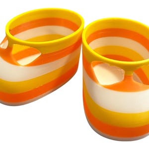 Może przedstawiać: Para bucików dla niemowląt w paski w kolorze żółtym, pomarańczowym i białym. Buty mają zaokrąglony nosek i wycięty detal z boku. Buty wykonane są z twardego tworzywa sztucznego.