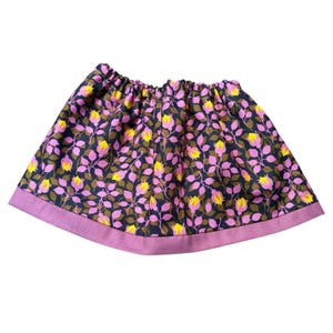 May include: A floral print skirt with an elastic waistband. The skirt features a dark navy background with pink, yellow, and brown floral patterns. The hem is a solid pink color. The skirt is made of a lightweight fabric.