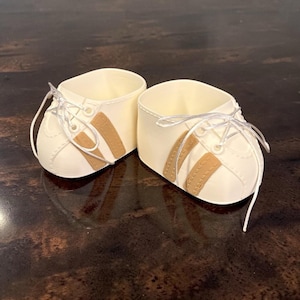 Peut inclure: Une paire de chaussures de poupée blanc cassé avec des bandes latérales beige, des lacets blancs et des détails cousus. Ces petites chaussures basses présentent un bout arrondi et une ouverture structurée, formant un ensemble assorti.