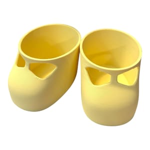 以下が含まれることがあります： 淡い黄色の3Dプリントのベビーシューズのペア。各靴は丸いつま先、切り欠きのあるストラップ、円筒形の本体を備えています。靴は無地のマットな素材でできており、メリージェーンスタイルの靴に似せてデザインされています。