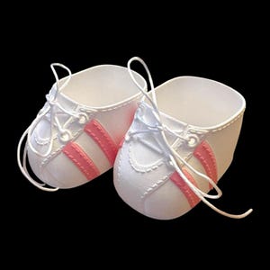 May include: A pair of white baby shoes with pink stripes and white laces. The shoes have a classic sneaker design, perfect for a newborn. The shoes are set against a black background.
