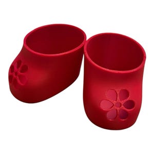 May include: A pair of bright red, small, open-topped shoes. Each shoe has a flower-shaped cutout on the front. The shoes are made of a solid material and are designed to fit a small object.