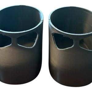 Peut inclure: Deux récipients cylindriques gris foncé au design unique. Chaque récipient a un large dessus ouvert et une découpe distinctive près du haut, ressemblant à un visage stylisé. Les récipients sont probablement imprimés en 3D, avec des lignes de couche visibles.