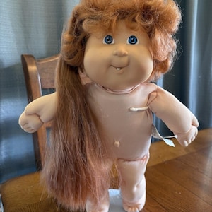 Może przedstawiać: Zabytkowa lalka Cabbage Patch Kid z brzoskwiniową skórą, niebieskimi oczami i rudymi włosami. Lalka ma miękkie ciało i nosi jasnoróżowy strój. Lalka stoi na białej podstawie.