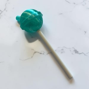 Op de afbeelding: Een turquoise lolly met een bolvormige snoepkop en een witte stok. Het snoepje heeft een textuur en staat op een witte marmeren achtergrond. De lolly is een enkel, geïsoleerd object.