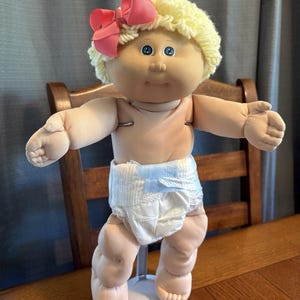 May include: A doll with light skin, blue eyes, and curly yellow yarn hair styled with a large pink ribbon bow. The doll has a soft fabric body, is wearing a white disposable nappy, and is supported by a white plastic stand on a wooden surface.