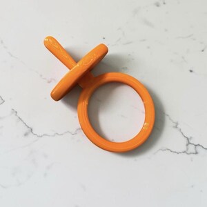 May include: A bright orange plastic dummy featuring a circular ring handle attached to a flat, disc-shaped shield and a short, straight teat component. The object has a smooth, matte finish and a uniform, solid orange colour throughout its structure.