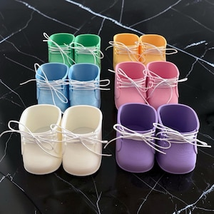 May include: A collection of twelve miniature shoes arranged in pairs, featuring colours including green, yellow, light blue, pink, white, and purple. Each shoe has a smooth finish, a rounded toe, and thin white laces tied in a bow at the front.