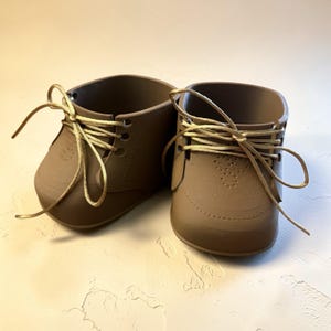 May include: Two brown decorative planters in the shape of baby shoes. The planters feature laces and a stitched design. The colour is a muted brown, with lighter tan laces. They are displayed on a white surface.