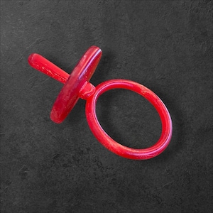 Puede incluir: Un chupete rojo, con un asa circular y una tetina. El chupete está hecho de un material translúcido. El objeto está sobre un fondo oscuro.