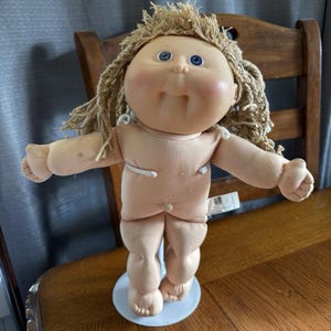 Könnte beinhalten: Eine Vintage-Puppe mit einem weichen, fleischfarbenen Körper, blauen Augen und blondem Garnhaar. Die Puppe steht auf einem hellblauen Ständer. Die Arme der Puppe sind ausgestreckt, und sie hat einen lächelnden Ausdruck.