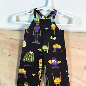 Op de afbeelding: Zwarte tuinbroek met een kleurrijke all-over print van diverse cartoonmonsters in tinten groen, paars, geel en oranje. Het kledingstuk heeft dunne schouderbanden die aan de voorzijde zijn bevestigd met felgroene ronde knopen.