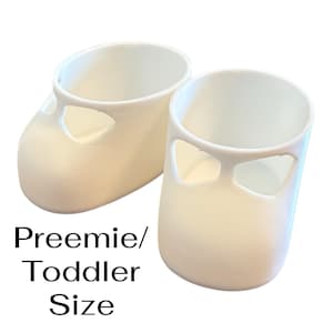 May include: Two off-white baby shoes with a cutout design on the sides. The shoes are made of a smooth material and are designed for preemie or toddler sizes. The text "Preemie/Toddler Size" is displayed below the shoes.