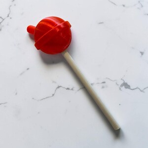 May include: A bright red lollipop with a round candy top and a light-colored wooden stick. The lollipop is set against a white marble background, creating a simple and clean aesthetic. The candy is a classic treat.
