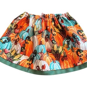 May include: A colorful skirt featuring a vibrant pumpkin pattern in shades of orange, teal, and white. The skirt has an elastic waistband and a green trim along the bottom edge, perfect for fall. The design includes pumpkins, gourds, and corn.