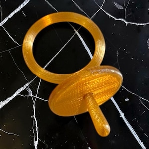 Könnte beinhalten: Ein durchscheinendes bernsteinfarbenes Objekt, bestehend aus einem kreisförmigen Ring und einer separaten kreisförmigen Scheibe mit einem zentralen zylindrischen Stift an der Unterseite. Die Oberfläche zeigt feine horizontale Fertigungslinien.