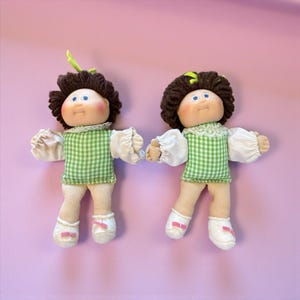 May include: Two vintage dolls with brown yarn hair, blue eyes, and rosy cheeks. They wear green and white checkered dresses with white lace collars and sleeves. The dolls have white socks with pink bows and are posed against a pink background.