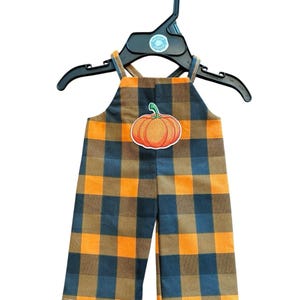 May include: Overalls with a pumpkin applique, hanging on a black hanger. The overalls feature a plaid pattern in orange, navy blue, and brown. The pumpkin is orange with a green stem. The overalls are designed for a baby or toddler.