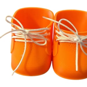May include: A pair of bright orange shoes with white laces tied in bows. The shoes have a rounded shape and a textured surface. The laces are neatly tied, creating a classic look. These shoes are likely for a baby or toddler.