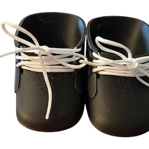 May include: A pair of black doll shoes with white laces. The shoes have a classic design, with a rounded toe and a lace-up closure. The laces are tied in a bow. The shoes are made of a smooth material.