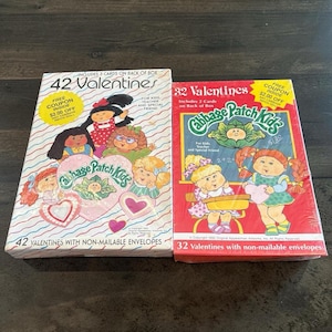 Può includere: Due scatole di biglietti di San Valentino Cabbage Patch Kids vintage. Una scatola è bianca con cuori rosa e la scritta "42 Valentines". L'altra scatola è rossa con la scritta "32 Valentines". Entrambe le scatole presentano illustrazioni di Cabbage Patch Kids e includono coupon.
