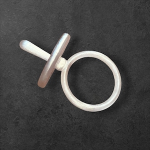 Puede incluir: Un anillo plateado con una banda circular y una barra horizontal que intersecta una barra vertical. El anillo tiene un acabado liso y pulido y está sobre un fondo oscuro y texturizado. El diseño es minimalista y moderno.